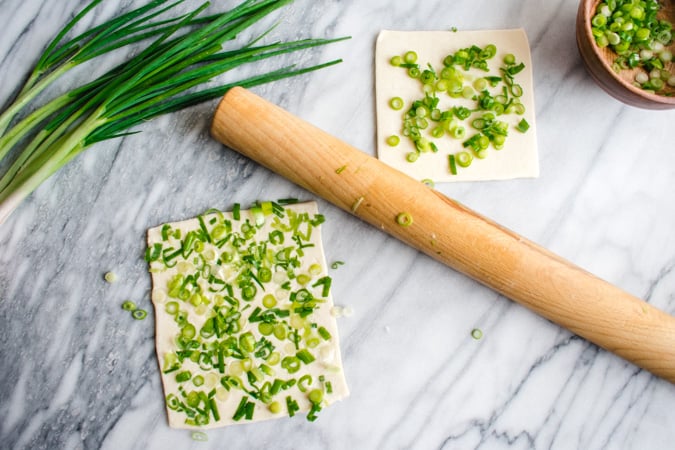 Chopped scallions on puff pastry squares with a rolling pin on a marble board
